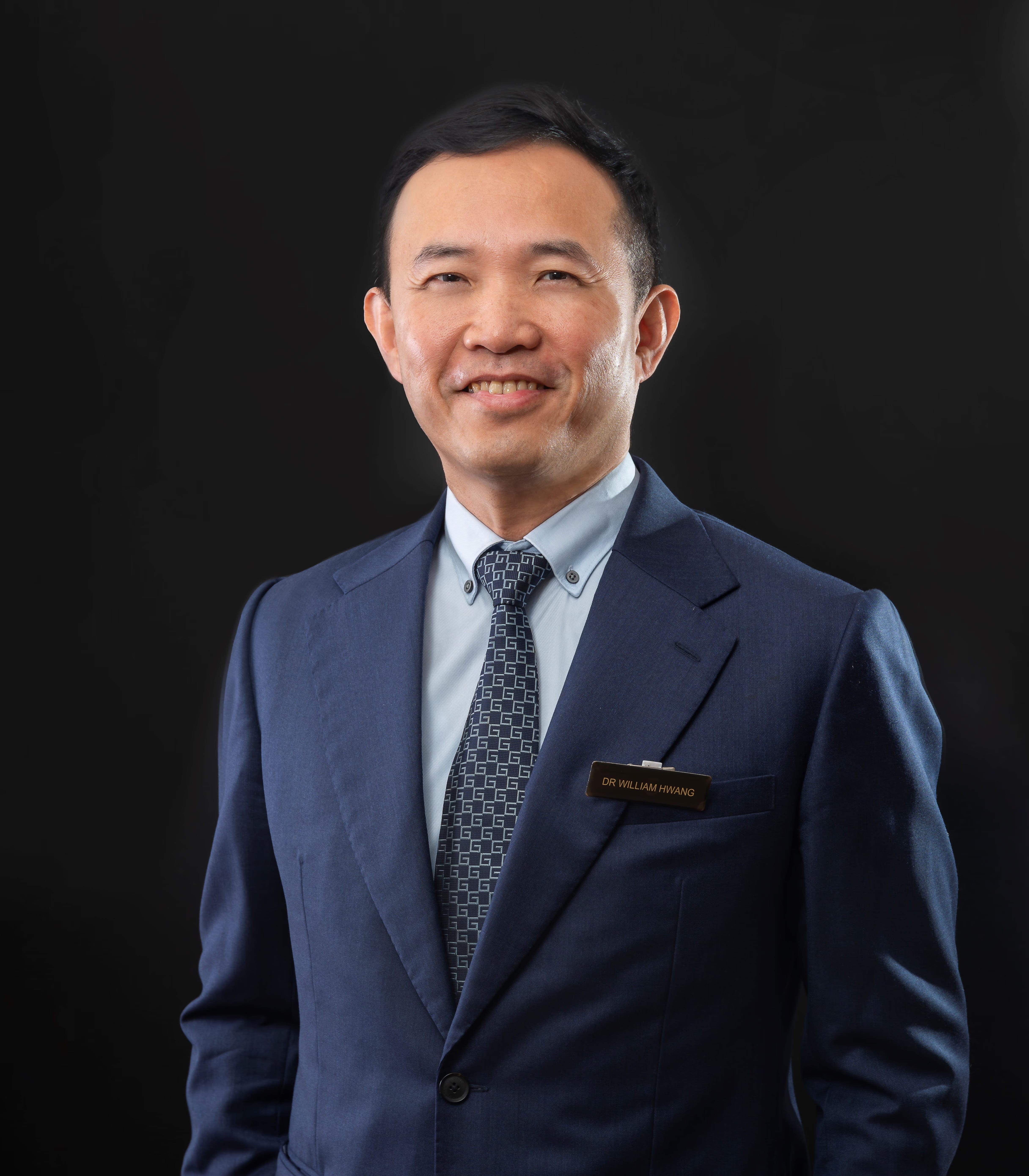 Man in a navy blue suit and patterned tie wearing a name badge reading Dr William Hwang, smiling against a dark background.